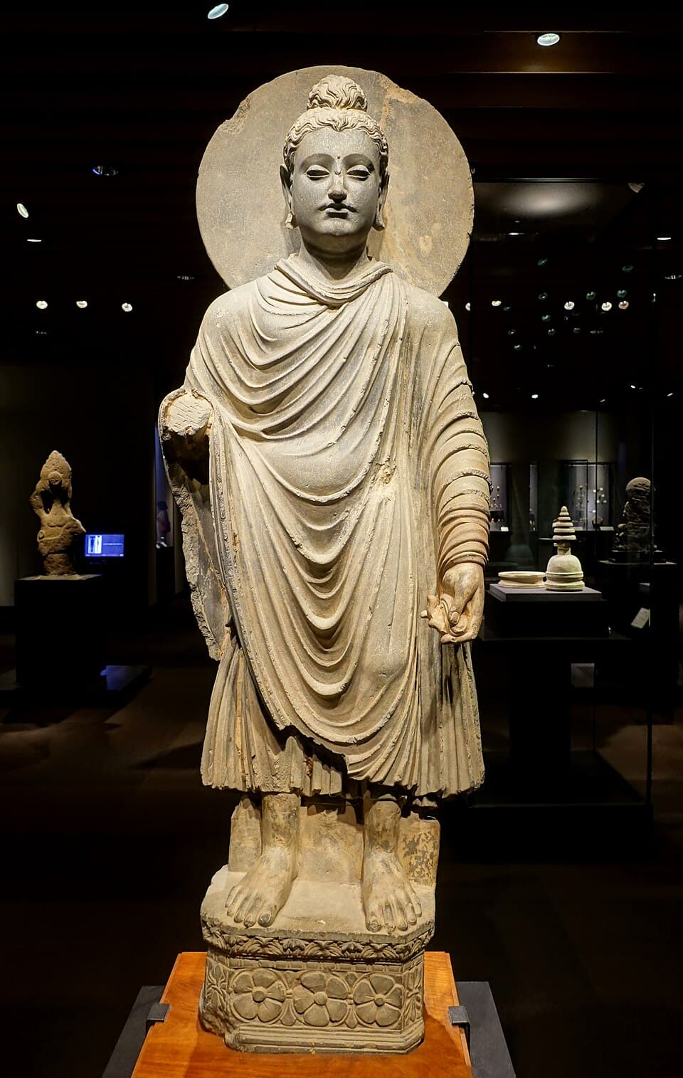 A Gandhara Buddha: The Gandhara school worked in grey blue schist in a strongly Hellenistic style, the Buddha with wavy hair and a toga like robe.